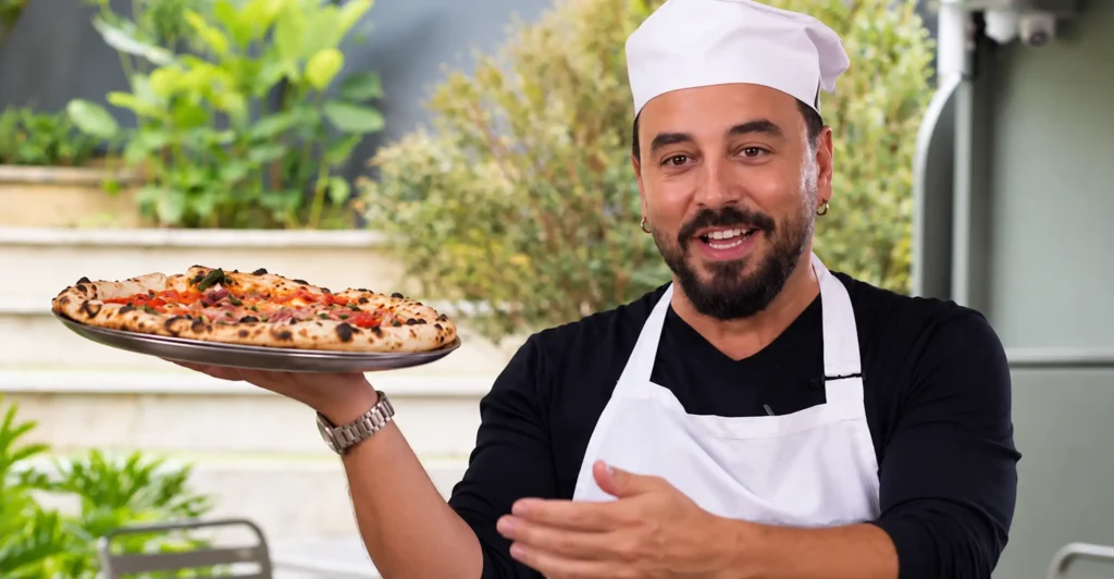 Tulio Recomienda, conocido como Tulio Zuloaga, presenta una pizza mientras viste uniforme de cocina.